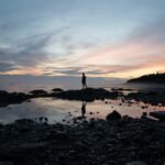 silhouette of person standing on rocky shore during sunset